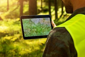 Person mit gelber Warnweste betrachtet im Wald eine topografische Karte auf einem Tablet